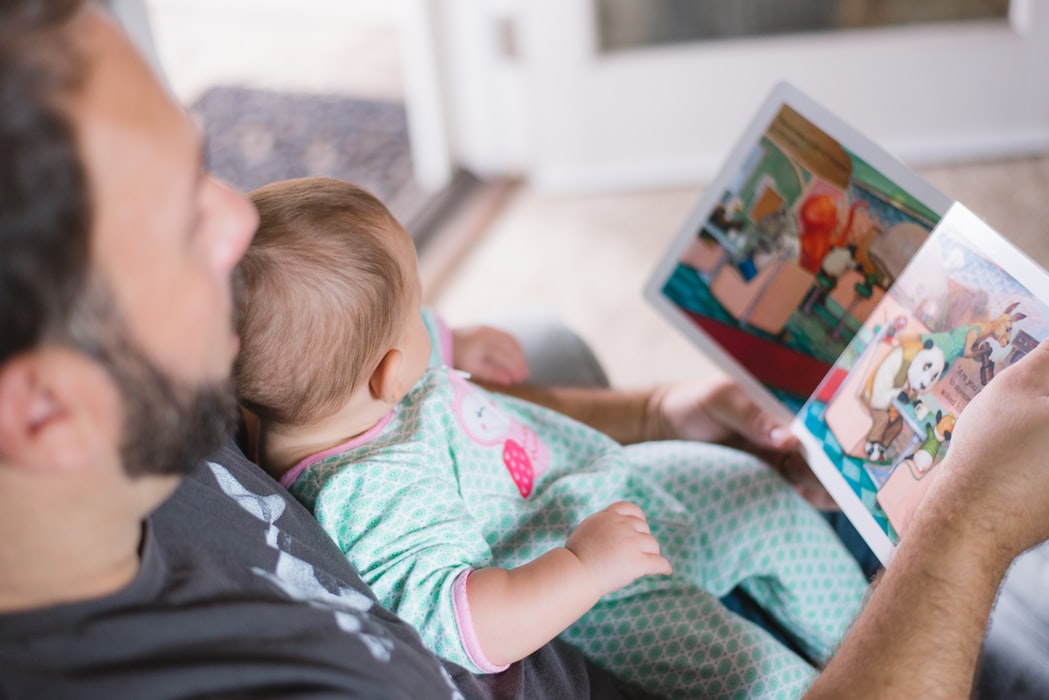A baby sits on their dads lap while he reads a book