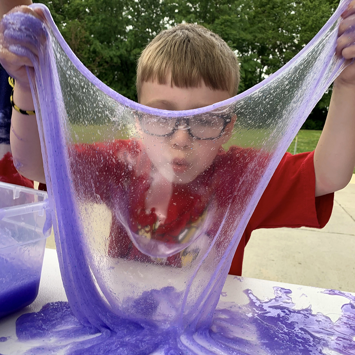 Child in a red shirt blows a bubble in purple slime outside