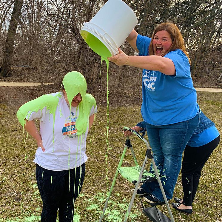 Woman in a blue shirt shows excitement at dumping green slime on a woman in a white shirt outside