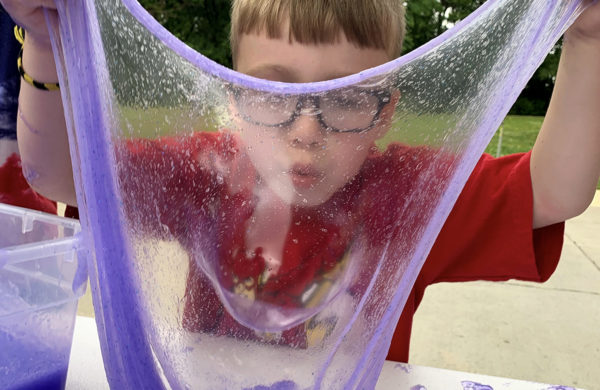 Child in a red shirt wearing glasses blows a bubble in purple slime